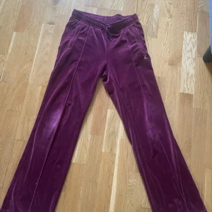 Vinröda velourbyxor från Juicy Couture - Mysiga vinröda velourbyxor från Juicy Couture med strassdetaljer och texten 'Juicy' på baksidan. Byxorna har normal passform och resår i midjan för extra komfort. Perfekta för en avslappnad och trendig look. Lite slitna vid knäna men annars fina!!