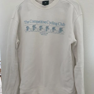 Vit sweatshirt från H&M med cykeltryck - Säljer en vit sweatshirt från H&M med blått tryck 'The Competitive Cycling Club' och cyklister på framsidan. Tröjan har relaxed fit och långa ärmar, perfekt för en avslappnad stil.