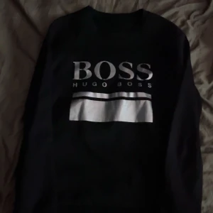 Svart sweatshirt från Hugo Boss - Säljer en svart sweatshirt från Hugo Boss med stor logga och text på bröstet i vitt och silver. Tröjan har rund halsringning och långa ärmar. Perfekt för en stilren och avslappnad look.