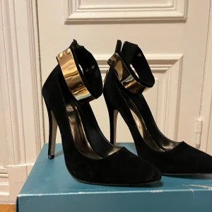Svarta pumps med guldfärgat ankelband - Säljer ett par  svarta pumps med spetsig tå och höga klackar. Skorna har ett brett, guldfärgat ankelband. Aldrig använd.