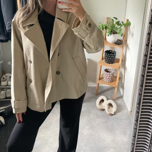 Beige kort trenchcoat med knappar - Säljer en beige kort trenchcoat med dubbelknäppning och bred krage. Aldrig använd!!