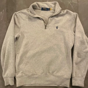 Grå tröja med half zip från Polo Ralph Lauren - Säljer en stilren grå tröja från Polo Ralph Lauren med halv dragkedja och broderad logga på bröstet. Tröjan har lång ärm och klassisk passform. Tröjan passar längre personer också.