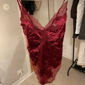 Vinröd satinlinne med spetsdetaljer - Säljer ett vinrött linne i satin med vackra spetsdetaljer längs urringning och nederkant. Linnet har smala axelband och en djup v-ringning, perfekt för en elegant look.