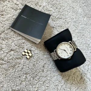 Silverfärgad klocka från Marc by Marc Jacobs - Stilren klocka från Marc by Marc Jacobs med silverfärgat armband i metall och vit urtavla. Klockan har tydliga bokstäver på urtavlan och en klassisk länkdesign. Levereras med originalask och extra länkbitar. Batteriet har varit urladdat ett tag men ska vara att bara byta 