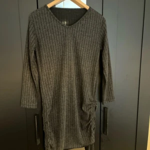 Mörkgrå ribbad klänning med v-ringning - Mysig mörkgrå klänning med ribbad struktur och v-ringad hals. Klänningen har trekvartsärm och en smickrande passform som passar perfekt till kyligare dagar.