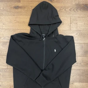 Svart hoodie med dragkedja från Polo Ralph Lauren - Svart hoodie från Polo Ralph Lauren med dragkedja framtill och klassisk logga broderad på bröstet. Tröjan har huva med snörning och två fickor framtill. Perfekt för en avslappnad stil. Använd fåtal gånger