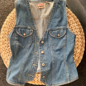 Jeansväst från Highway - Säljer en klassisk blå jeansväst från Highway i 100% bomull. Västen har knappar framtill, två bröstfickor med lock och knapp samt ett justerbart bälte i ryggen. 