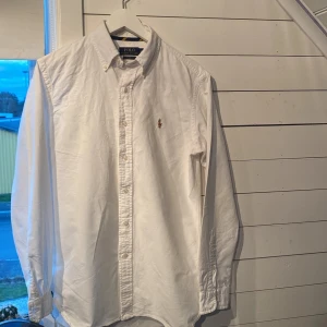 Vit skjorta från Polo Ralph Lauren - Säljer en klassisk vit skjorta från Polo Ralph Lauren med broderad logga på bröstet. Skjortan har långa ärmar och knappar framtill. Perfekt till sommaren!! Skjortan är i bra skick. Billligare vid snabb affär!                                                 Priset är pga ett litet håll vid en av knapparna som då är lagat och syns ej när skjortan är knäppt. (Lagning bild 5)