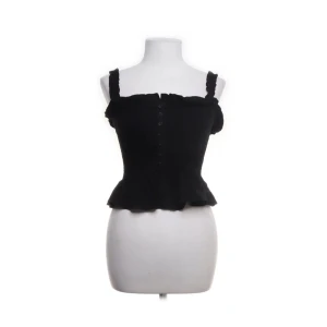 Svart croppad topp med volang - Säljer en svart croppad topp med volangkant nertill och axelband med rynkade detaljer. Toppen har knappar framtill och en feminin siluett, perfekt för att lyfta din outfit.