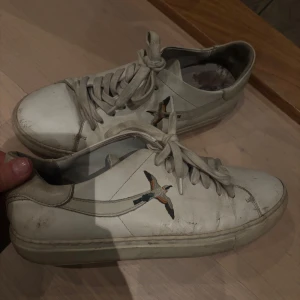 Vita sneakers från Axel Arigato med broderad fågel - Säljer ett par vita sneakers från Axel Arigato med broderad fågel på sidan och guldfärgad logga. Skorna har snörning och platt sula, klassisk rund tå och är tillverkade i skinn. Perfekta för dig som gillar stilrena och trendiga sneakers.