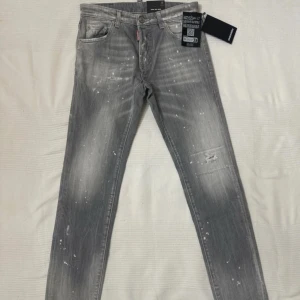 Grå jeansbyxor från Dsquared2 Cool Guy Jean - Säljer ett par grå Dsquared2 Cool Guy Jean med slitningar och vita färgstänk. Byxorna har fem fickor, knappgylf och en smal passform. Märkeslapp och etiketter sitter kvar. Perfekta för dig som gillar en edgy streetstil. Om ni vill ha flera bilder DM mig!