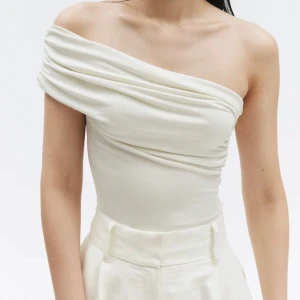 Vit oneshoulder bodysuit - Säljer en stilren vit bodysuit med draperad design och oneshoulder-skärning. Toppen har en elegant och asymmetrisk form som ger en modern look. Perfekt att styla med både byxor och kjol. Lappen sitter kvar, alltså helt oanvänd.