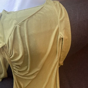 Ljusgrön omlottklänning - Säljer en ljusgrön klänning med omlottdesign och lätt glansigt material. Klänningen har lång ärm och snygga rynkade detaljer längs sidan. Och slit. Perfekt för dig som vill ha något stilrent men ändå unikt. Den är lite stretchig. Använd 1 gång 