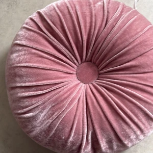 Rund rosa sammetskudde - Säljer en rund kudde i mjuk rosa sammet med dekorativa veck och en knapp i mitten. Kudden har en lyxig känsla och passar perfekt som inredningsdetalj i soffan eller på sängen.