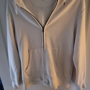 Vit zip hoodie från Uniqlo - Säljer en stilren vit hoodie från Uniqlo i storlek M för jag har växt ur den (jag är 190cm lång och väger 78 kg). Tröjan har dragkedja framtill, två fickor och huva med snörning. Perfekt för en avslappnad look och enkel att matcha med andra plagg.