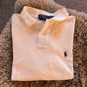 Polo Ralph Lauren Piké - Säljer en vit Polo Ralph Lauren piké med en krage och knappar. Pikén har en liten flävk som man knappt kan se. Tröjan har även liten reva längst ner i slitsen. Perfekt för sommaren.
