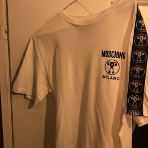 Vit t-shirt från Moschino Milano - Säljer en vit t-shirt från Moschino Milano med svart tryck på bröstet och ett brett band med Moschino-loggor längs ena sidan. Klassisk passform och rund halsringning. Perfekt statement-plagg för dig som gillar designerkläder.