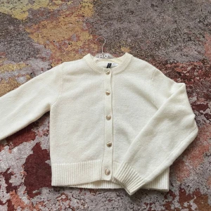 Vit kofta med knappar från H&M - Mysig vit kofta med rund halsringning och snygga guldfärgade knappar framtill. Tröjan har långa ärmar och breda ribbade muddar vid ärmslut och nederkant. Perfekt för lager-på-lager och enkel att matcha.