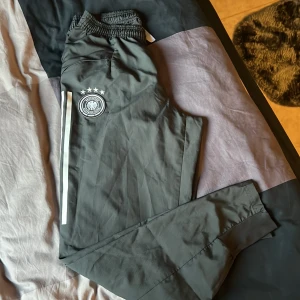 Svarta träningsbyxor från Adidas med Tysklands landslagslogga - Säljer ett par svarta träningsbyxor från Adidas med Tysklands landslagslogga och fyra stjärnor på ena benet. Byxorna har vita ränder längs sidorna och resår i midjan. Perfekta för fotbollsträning eller sportiga dagar.