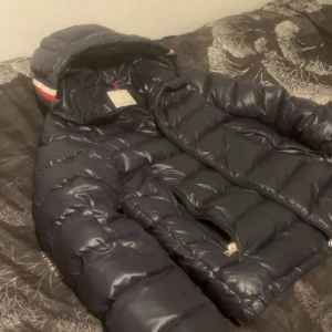Svart pufferjacka med huva från Moncler - Säljer en svart pufferjacka från Moncler med luva och dragkedja. Jackan har långa ärmar och detaljer i rött och vitt vid ärmslut och luva. Perfekt för kyliga dagar och riktigt snygg streetstil. Mycket bra skick, hör av dig vid funderingar!
