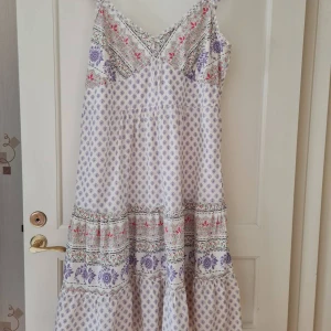 Sommarklänning med blommigt mönster från KappAhl strl 48 - Säljer en långklänning i vitt med lila och rosa blommigt mönster. Klänningen har smala axelband, v-ringning fram och rynkade detaljer upptill. Perfekt för sommarens varma dagar eller midsommarfirande. 100% bomull. Etiketten är klippt. Storlek 48. Kan hämtas i Vallentuna.