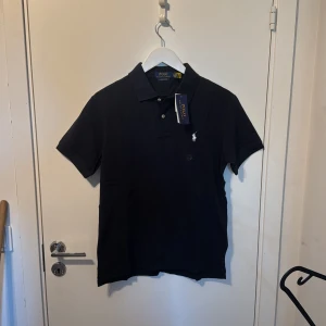 Mörkblå pikétröja från Polo Ralph Lauren - Säljer en svart pikétröja från Polo Ralph Lauren i modellen Custom Slim Fit. Tröjan har korta ärmar, klassisk krage och två knappar framtill. Ikoniska broderade loggan i vitt på bröstet. Perfekt för en stilren look.