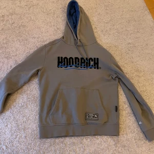 Grå hoodie från Hoodrich - Säljer en grå hoodie från Hoodrich med svart och blå logga på bröstet och ryggen. Tröjan har huva, magficka och en patch nertill. Perfekt för en avslappnad stil. Knappast använd.✨