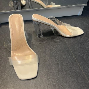 Vita klackskor med transparent rem och klack - Säljer ett par trendiga vita klackskor med fyrkantig tå och transparenta remmar samt klack. Perfekta för dig som vill ha en modern och stilren look med en twist. Klacken är också transparent för extra effekt.
