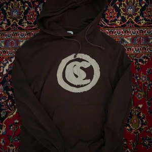 Brun hoodie från COLLUSION med tryck - Säljer en brun hoodie från COLLUSION med en stor beige cirkel och grafiskt tryck på bröstet. Tröjan har huva med dragsko och en klassisk känguruficka framtill. Perfekt för en avslappnad stil.