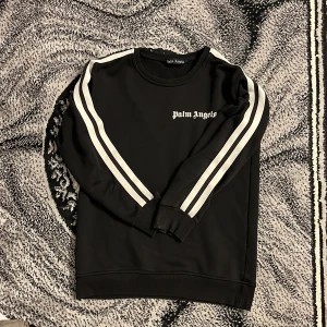 Svart sweatshirt från Palm Angels med vita ränder - Svart sweatshirt från Palm Angels med vita ränder längs ärmarna och logga på bröstet. Tröjan har rund halsringning och långa ärmar. Perfekt för en sportig och stilren look.