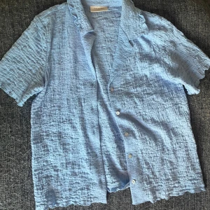 Ljusblå krinklad blus med volangkrage - Säljer en ljusblå skjorta från free/quent med korta ärmar och volangdetalj vid kragen. Skjortan har knappar framtill och är varsamt använd. Jättesöt till sommarn/vår!