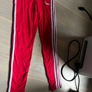 Röda Adidas leggings med vita ränder för barn - Säljer ett par röda Adidas leggings med klassiska vita ränder längs sidorna och Adidas-logga på höften. Tight passform och elastisk midja, perfekta för träning eller en sportig look. Dom är är för barn mellan 9-11 år eller 134-146