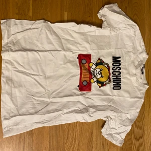 Vit t-shirt med nalletryck från Moschino - Vit t-shirt från Moschino med stort tryck av en nallebjörn i färgglada detaljer och texten MOSCHINO på bröstet. Klassisk passform och rund halsringning. Perfekt statement-plagg för dig som gillar lekfull stil.