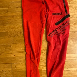 Röd hoodie och byxor från Hoodrich - Säljer ett rött set från Hoodrich med svart tryck och dragkedjor. Hoodien har huva och hel dragkedja framtill, samt fickor. Byxorna har resår i midjan och svarta detaljer. Perfekt för en avslappnad stil.