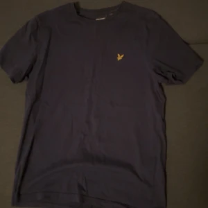 Mörkblå t-shirt från Lyle & Scott - Säljer en mörkblå t-shirt från Lyle & Scott med klassisk passform och korta ärmar. T-shirten har en liten gul broderad logga på bröstet. Perfekt till vardags och enkel att matcha med andra plagg.