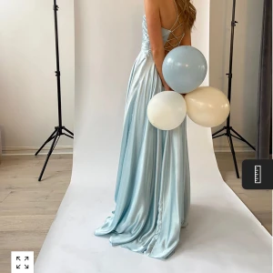 Ljusblå långklänning med omlott och smala axelband - Säljer en elegant ljusblå långklänning med omlottliv och smala axelband. Klänningen har öppen rygg med snörning och ett vackert, glansigt satintyg som faller fint. Perfekt för bal!