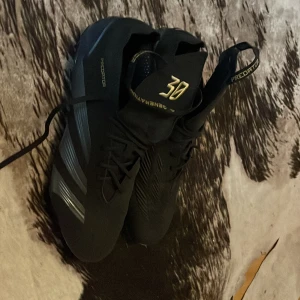 Svarta Adidas Predator fotbollsskor - Säljer ett par svarta Adidas Predator fotbollsskor med guldiga detaljer och nummer 30 på plösen. Skorna har snörning och är designade för bra grepp på planen. Perfekta för dig som vill sticka ut på matchen!