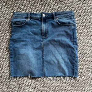 Blå jeanskjol från Pieces - Säljer en klassisk blå jeanskjol från Pieces i storlek XL. Kjolen har rå kant nertill, fem fickor och bälteshällor. Materialet är en blandning av bomull och polyester, vilket gör den både mjuk och slitstark. Perfekt till sommaren!
