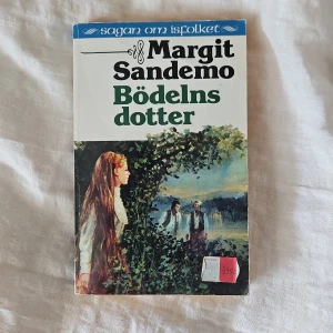 Bödelns dotter - Säljer boken 'Bödelns dotter' av Margit Sandemo, del av serien Sagan om Isfolket. Omslaget har en illustration med en kvinna med långt rött hår och flera personer i bakgrunden. Boken är på svenska och utgiven av Boknöje AB.