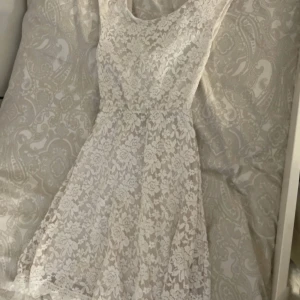 Vit spetsklänning med öppen rygg - Superfin vit spetsklänning med ärmlös design och öppen rygg. Klänningen har ett vackert blommigt spetsmönster och en lätt utställd kjol. Perfekt för sommarens speciella tillfällen eller studentfirande.