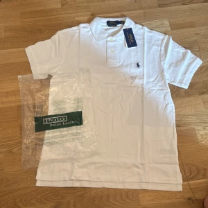 Vit pikétröja från Polo Ralph Lauren - Klassisk vit pikétröja från Polo Ralph Lauren med broderad logga på bröstet. Tröjan har krage, knappar vid halsen och är kortärmad. Perfekt för en stilren och tidlös look.