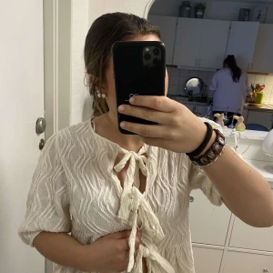 Vit strukturerad blus med knyt från Gina Tricot - Supersöt vit blus från Gina Tricot med strukturerat tyg och två knyt framtill. Blusen har korta puffärmar och en lätt volang nedtill. Perfekt för vår och sommar, och ger en romantisk vibe.