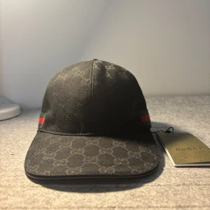 Svart keps från Gucci  - Skriv privat för video. Svart keps från Gucci med diskret GG-monogram över hela ytan. Kepsen har klassisk böjd skärm och justerbar rem baktill. På sidorna finns gröna och röda ränder som signaturdetalj. Märkets logotyp syns subtilt i tyget. Storlek ”M”