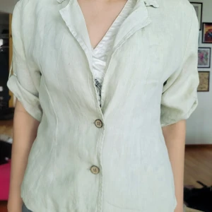 Ljusbeige linneskjorta / kavaj med kort ärm - Säljer en ljusbeige kavaj / skjorta i linne med korta ärmar och två knappar framtill. Ärmarna kan rullas upp och fästas med knapp. Färgen är svår att få rätt på bilderna, den har lite gult / grönt i sig. Tjejen på bilden har oftast storlek 40, ibland 38. Den r runt 46 cm från armhåla till armhåla och runt 54 cm lång. 