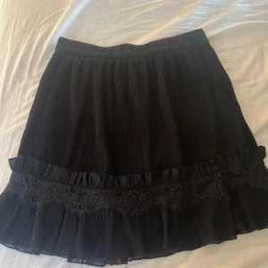 Svart kjol - Säljer en svart kortkjol med plisserat tyg och volangkant. Kjolen har en bred spetsdetalj och volang nertill som ger en söt och trendig look. Perfekt att styla med en enkel topp eller stickad tröja.