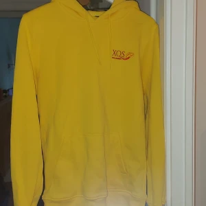 Gul hoodie med XQS-tryck - Säljer en gul hoodie från XQS med liten logga och tryck på bröstet. Tröjan har huva, magficka och långa ärmar. Perfekt för dig som gillar färg och vill ha en bekväm och stilren hoodie.
