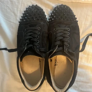 Svarta sneakers med nitar från Christian Louboutin - Säljer ett par svarta sneakers från Christian Louboutin med svarta nitar över hela tån och ovandelen. Skorna har klassisk röd sula och är tillverkade i mocka med snörning. Perfekt för dig som vill sticka ut med en edgy stil. Innersula saknas och 3 spikar