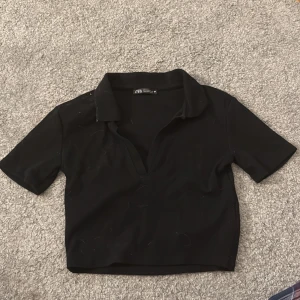 Svart croppad polotopp från Zara - Säljer en svart croppad polotopp från Zara. Toppen har korta ärmar, krage och djup v-ringning. Perfekt för en trendig och stilren look.