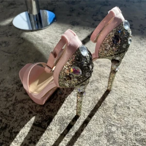 Glittriga rosa klackskor med stenar - Säljer ett par höga klackskor i ljusrosa med glittriga detaljer och stora dekorativa stenar. Skorna har öppen tå och rem runt vristen. Perfekta för dig som vill sticka ut och glänsa på festen!