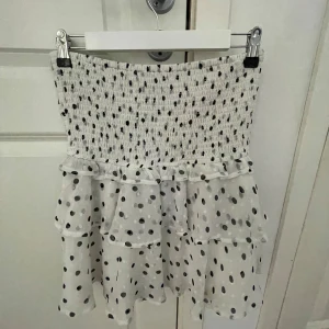 Vit prickig volangkjol från Lindex - Supersöt vit kjol med svarta prickar och volangdetaljer. Kjolen har smock i midjan för en bekväm passform och är kort i modellen. Säljer pga den är för stor på mig men sitter jätte fint på, pris går att diskutera och fråga för fler bilder!!